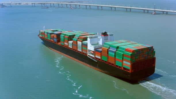 Container ship loaded with colorful containers sailing under bridge, representing global trade and drayage connection.