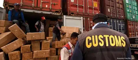 Worker performing customs clearance checks with clipboard in front of shipping containers and forklift