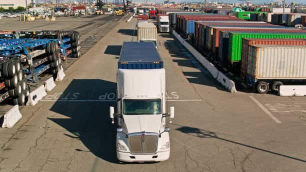 White drayage truck carrying container in port yard, showing short distance transport between terminal and warehouse.