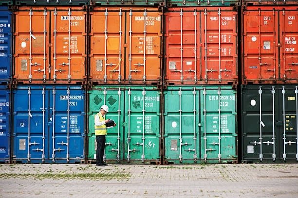 Worker performing safety inspection on stacked shipping containers for ground shipping.