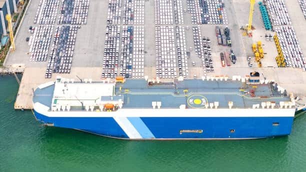 Roll on roll off cargo vessel docked with cars lined up on terminal, showing vehicle shipping and related drayage activities.