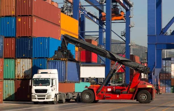 Red reach stacker loading a blue shipping container onto a white transport truck.