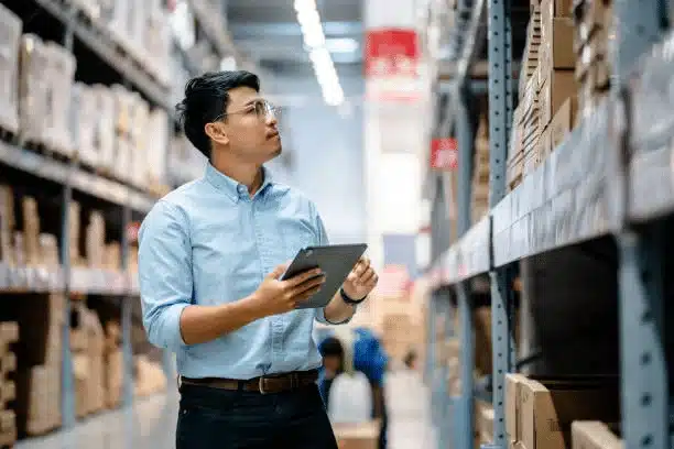 Fulfillment center worker checking stock with tablet in warehouse aisle.