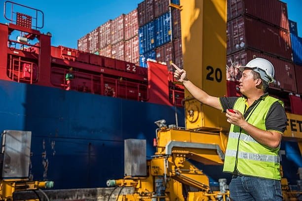 Worker in safety vest supervising cargo container loading onto a vessel.