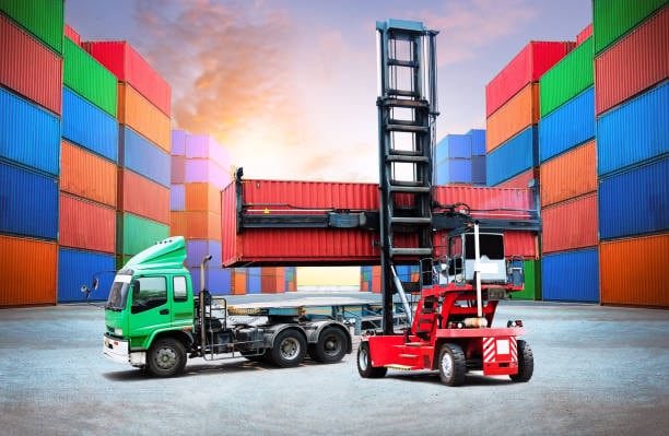 Reach stacker transferring a shipping container onto a truck surrounded by stacked cargo containers.