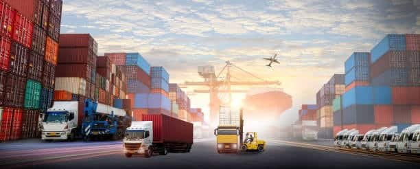 Large freight yard with shipping containers, trucks, cranes, and an airplane flying above.