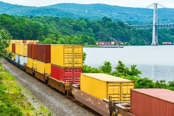 Freight train loaded with shipping containers moving along a river as part of ground shipping.