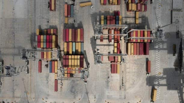 Top-down view of container stacks and cranes in a freight yard connected to global shipping channels.