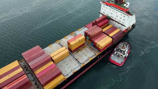 Container ship moving through a maritime shipping channel with tugboat assistance.