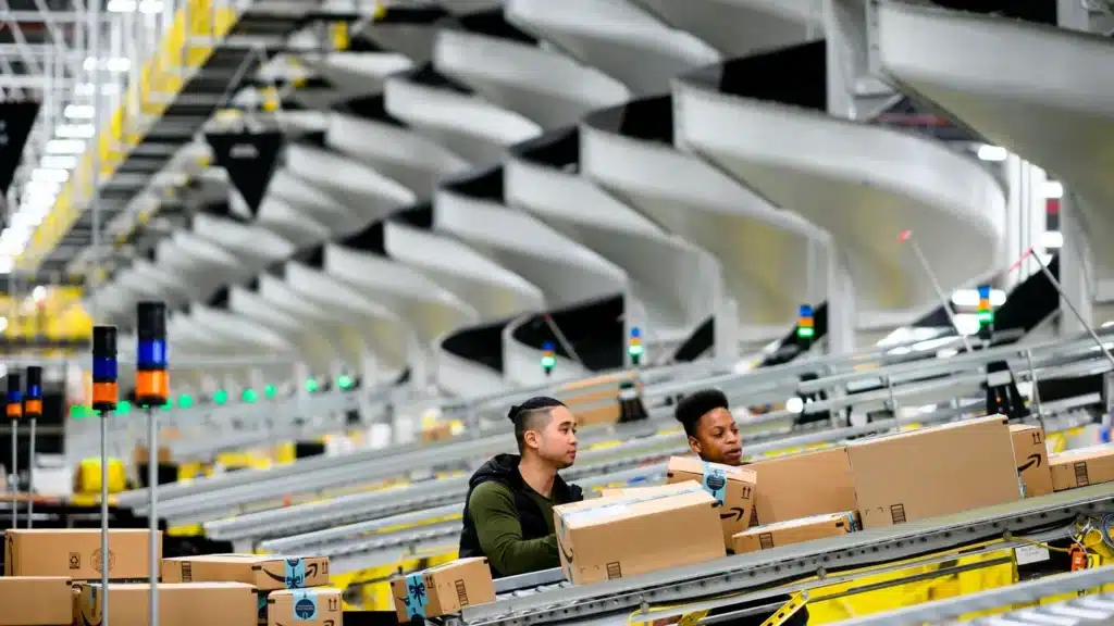 Workers sorting Amazon parcels on automated conveyor lines inside a large fulfillment center, showing the scale and efficiency of Amazon logistics operations.