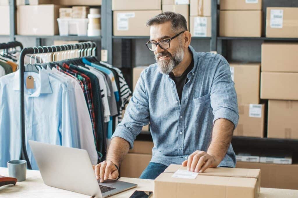 Business owner managing online apparel orders using laptop in a DTC fulfillment warehouse.