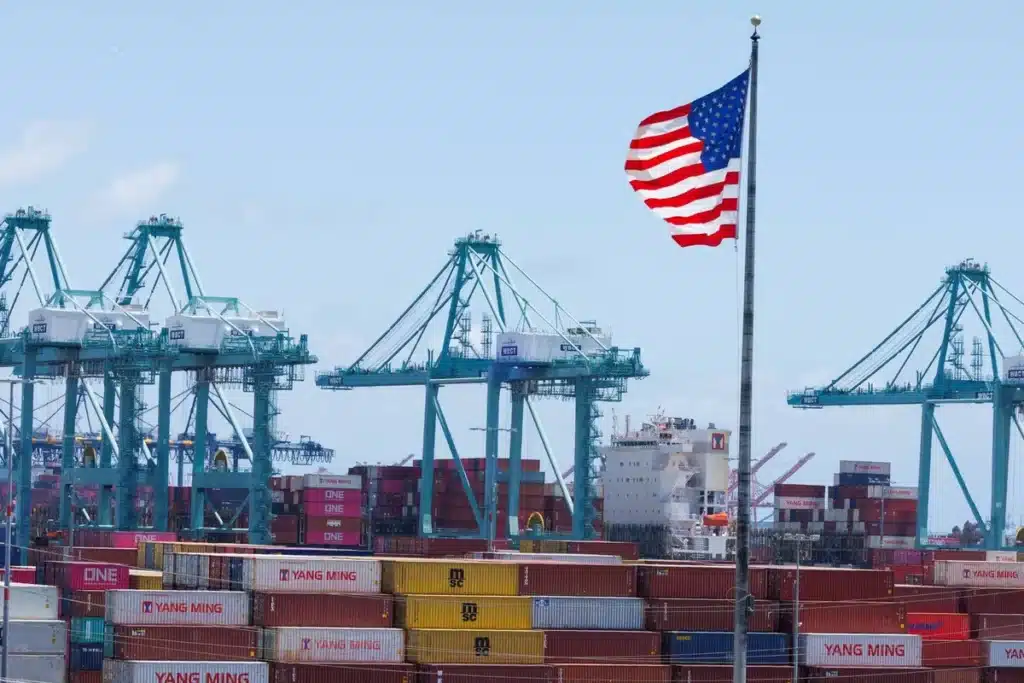 A U.S. port with cranes, shipping containers, and an American flag, illustrating the real cost of importing to the U.S. in 2025, including duties and taxes.