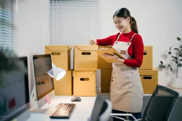 Woman checking AliExpress order list with shipping boxes in background.