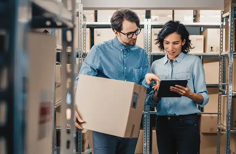 Warehouse staff checking inventory with a tablet while handling cartons, demonstrating efficient stock control in B2B fulfillment operations.