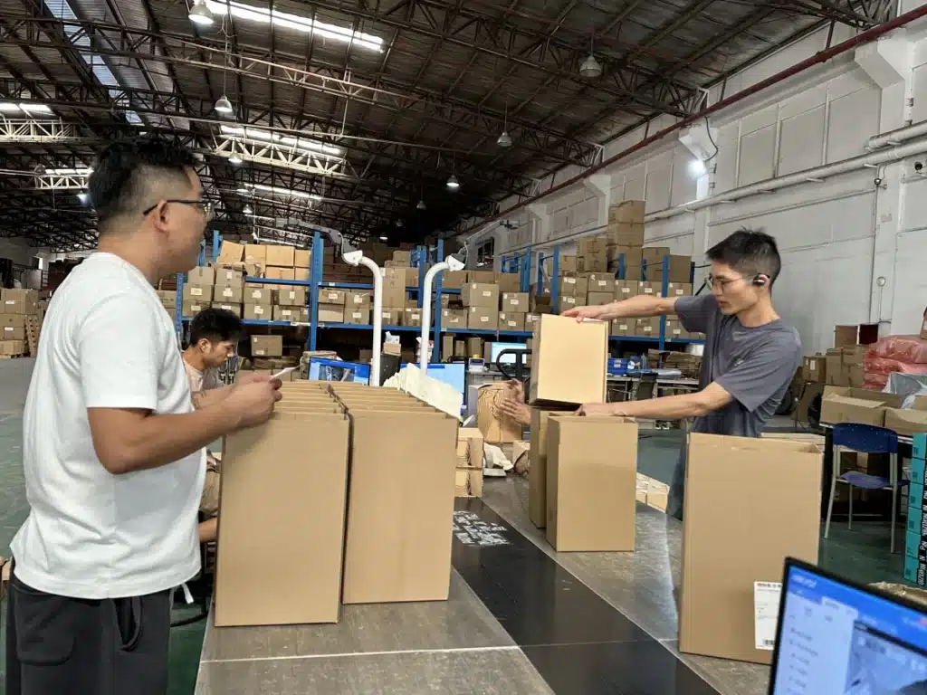 Warehouse staff packing shoeboxes into shipping cartons at workstations in a China 3PL fulfillment center.