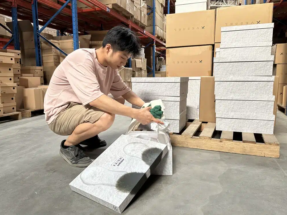Warehouse worker examining footwear and tissue packaging in an open shoebox beside palletized inventory