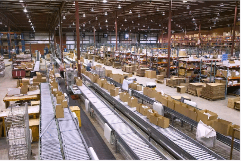 Workers sorting packages on a complex conveyor belt system in a busy distribution center, highlighting pick and pack fulfillment processes.
