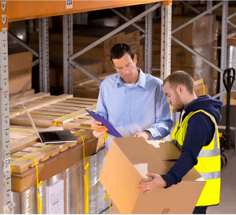 Two men in a warehouse, one with a laptop and clipboard, discussing inventory and packaging strategies with a worker holding a box.