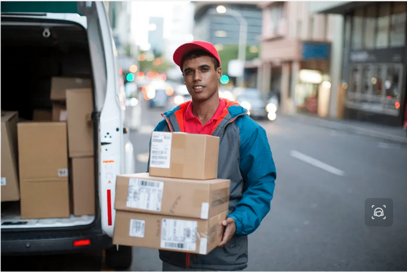 Smiling delivery driver holding stacked packages beside an open van on city street, symbolizing flexible last-mile solutions used to maintain on-time delivery during flash sale spikes.