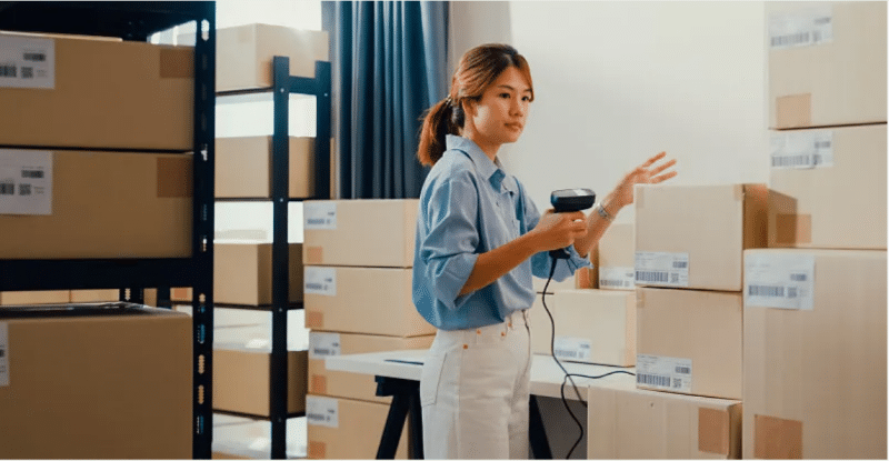 Female warehouse worker scanning barcodes on boxes while reviewing inventory records, demonstrating proactive management to prevent long-term storage fee accumulation.