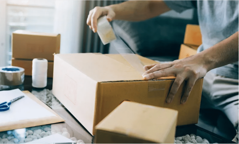 Person sealing a cardboard box with tape, preparing packages for shipping during the peak holiday season to minimize dimensional weight and handling fees.