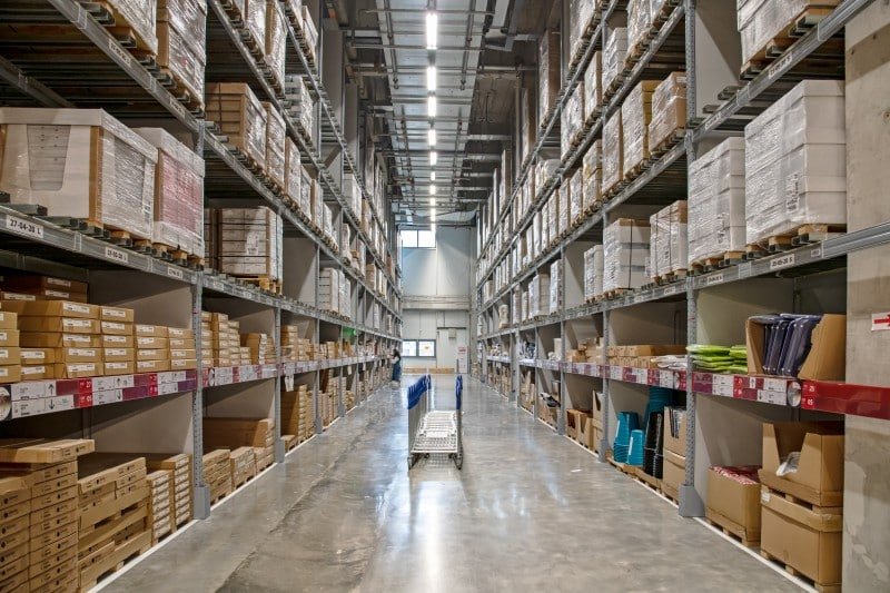 Narrow warehouse aisle with metal shelving units holding neatly arranged product bins and small parcels, featuring a manual cart for pickers — ideal for assembling customized subscription kits with minimal error rates.