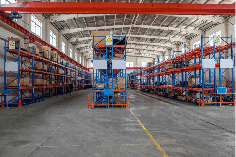 Wide-angle view of a modern distribution center with tall blue-and-orange pallet racking systems, overhead cranes, and organized aisles — designed to support high-volume subscription box assembly and recurring shipment cycles.