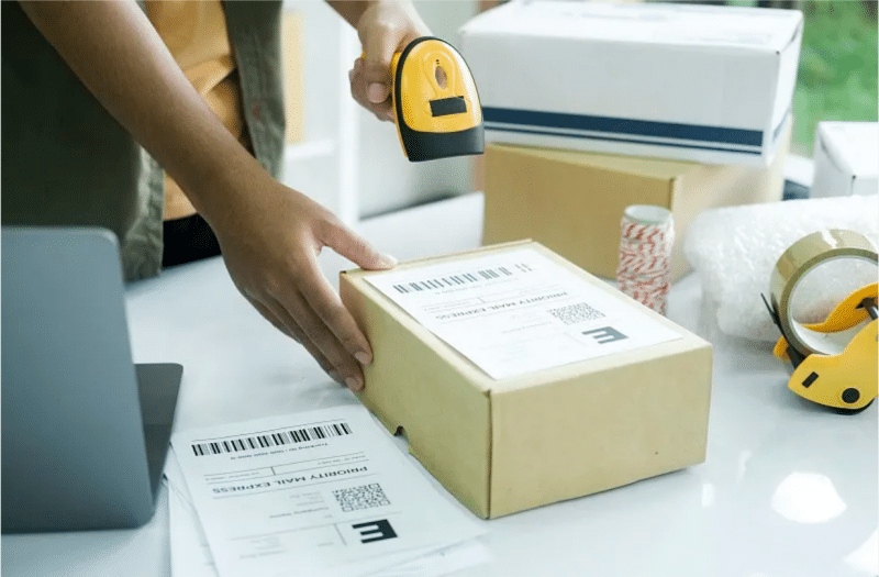 Person scanning barcode on a cardboard box labeled with shipping information, representing pre-order fulfillment workflow in e-commerce warehouse operations.