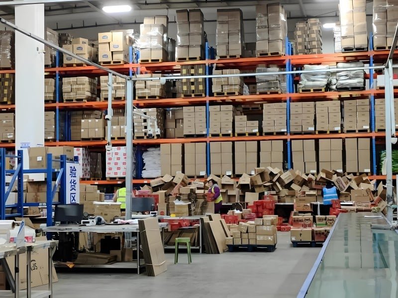 Busy fulfillment center packing station with stacks of cardboard boxes, packing materials, and workers verifying orders, highlighting the operational complexity and value-added services involved in shipping from China.